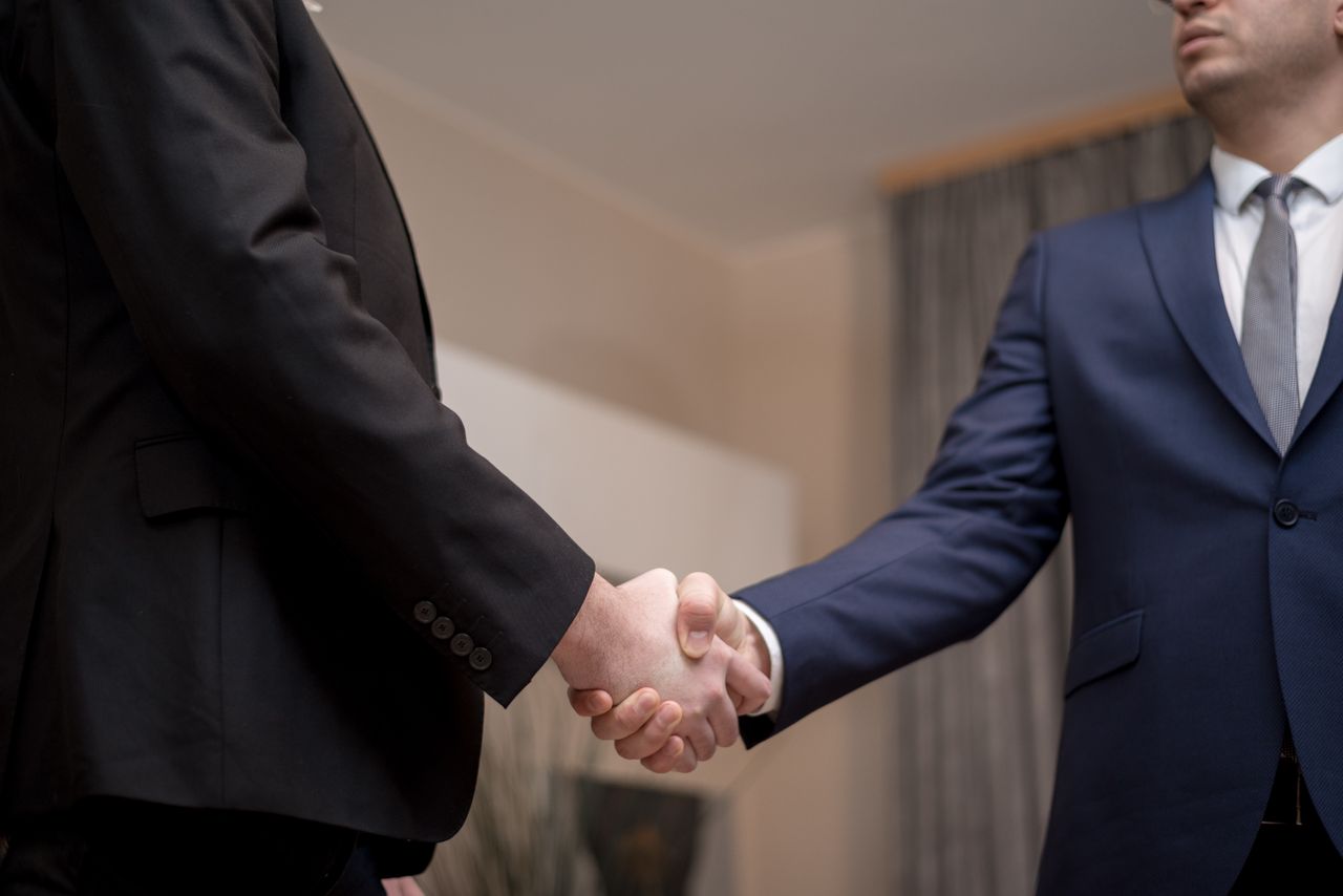 services-04 Two businessmen in suits shaking hands, emphasizing trust and partnership indoors.